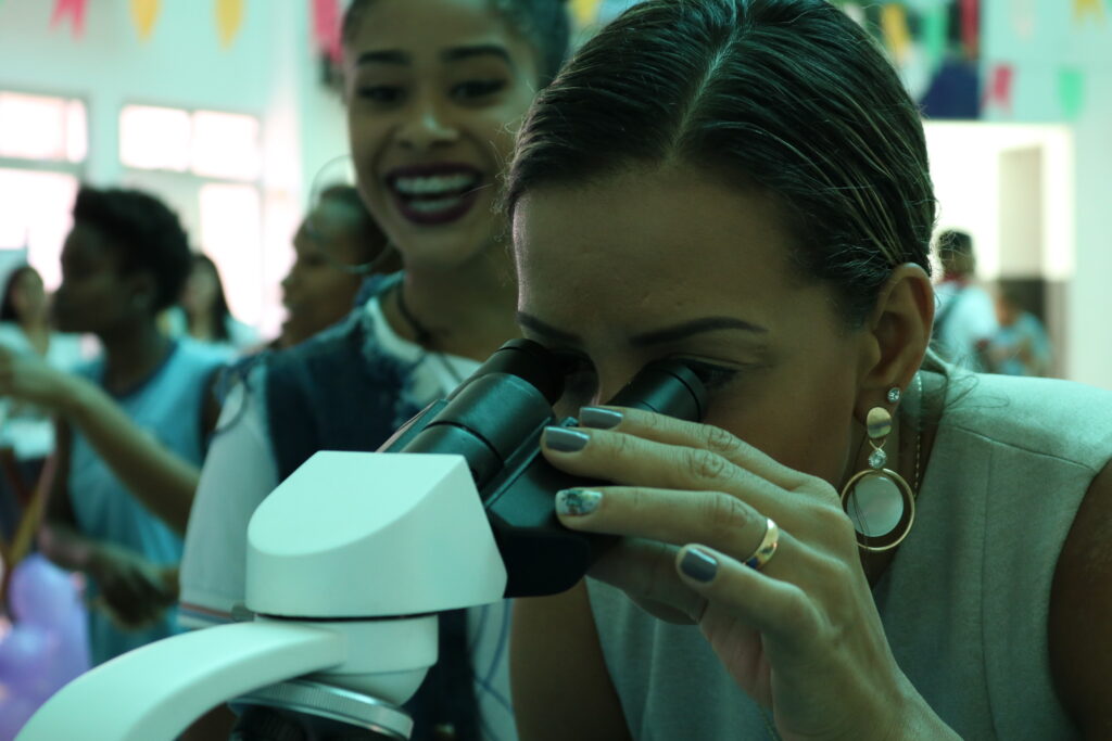 Mujer joven mirando a través de un microscopio. Otra joven, a su lado, la mira sonriendo.