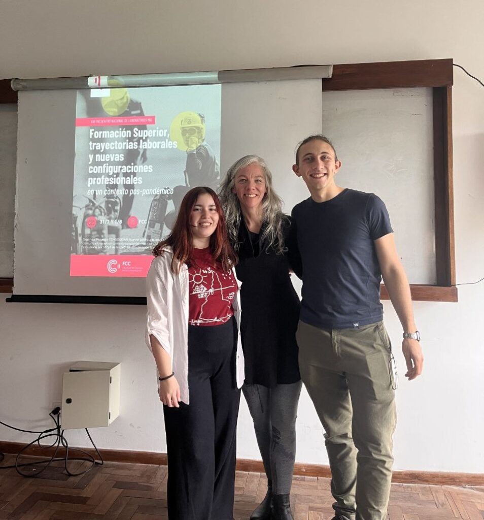 Tres personas de pie en un aula. De fondo hay un pizarrón donde está proyectado el afiche del evento.